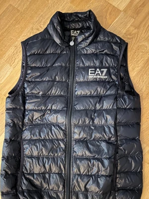 Svart dunväst från EA7 Emporio Armani - Snygg svart dunväst från EA7 Emporio Armani med quiltad design och hög krage. Västen har dragkedja framtill och logga på bröstet. Perfekt för lager-på-lager och streetstyle. Tillverkad i polyester och polyamid. Storlek L men är som en M i storleken. 