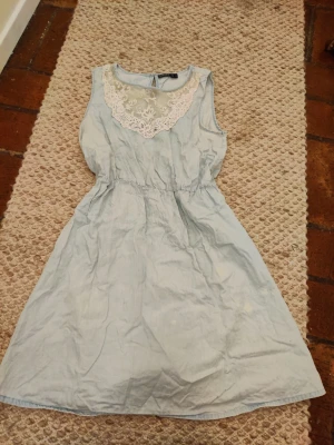 Ljusblå ärmlös klänning med spets - Söt ljusblå klänning med ärmlös design och vit spetsdetalj vid halsringningen. Klänningen har en markerad midja och lätt utställd kjol, perfekt för varma dagar. Tillverkad i bomull för en skön och luftig känsla.