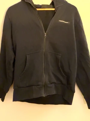 Svart zip-hoodie från Peak Performance - Klassisk svart hoodie med dragkedja från Peak Performance. Tröjan har två stora fickor framtill och diskret logga på bröstet. Tillverkad i mjukt bomullsmaterial och har ribbade muddar vid ärmslut och nederkant. Perfekt för en avslappnad och stilren look.