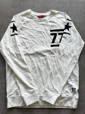 Vit sweatshirt med svarta detaljer - L - Vit långärmad sweatshirt från Lager157 med svarta tryck, inklusive siffran 77 på bröstet och stjärnor på axlarna. Rund halsringning och ribbade muddar vid ärmslut och nederkant. Perfekt för en avslappnad och cool look.