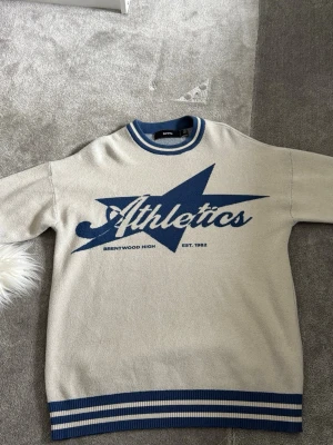 Beige college-tröja Athletics - Snygg beige college-tröja från Gorilla Wear med blåa detaljer och tryck 'Athletics' samt stjärna på bröstet. Ribbstickade muddar och krage i blått och vitt. Perfekt för en sportig och avslappnad stil.