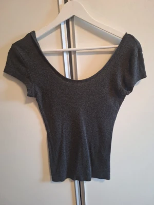 Mörkgrå ribbad topp med djup rygg - Snygg mörkgrå ribbad topp med korta ärmar och djup, rundad rygg. Figurnära passform och stretchigt material som sitter skönt på kroppen. Perfekt att matcha med jeans eller kjol för en trendig look