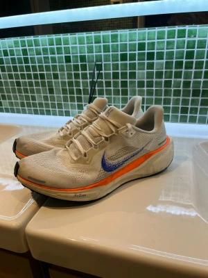 Nike ZoomX Vita sneakers - Nike ZoomX sneakers i vitt mesh med blå swoosh och orange detalj längs sulan. Lätta och ventilerande med sportig design och snörning. Sulan har Air Zoom-dämpning för extra komfort. Perfekta för dig som gillar stilrena och funktionella sneakers.