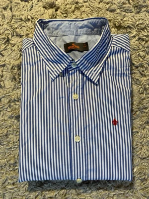 Blårandig skjorta från Morris - Klassisk blårandig skjorta från morris med vit botten och smala blå ränder. Skjortan har lång ärm, knappar framtill och en liten röd broderad logga på bröstet. Tillverkad i bomull för en skön och luftig känsla.