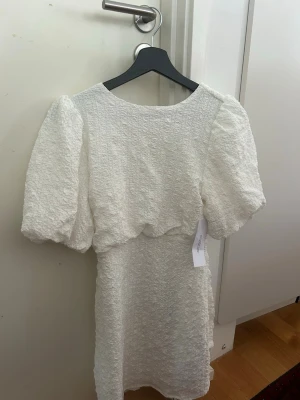 Vit kort klänning med puffärm och rosett - Supersöt vit kort klänning med puffärmar. Klänningen har öppen rygg med stor rosett. Perfekt som studentklänning!💕Oanvänd med lapparna kvar. 