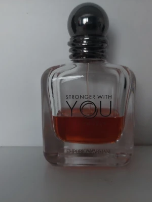 Emporio Armani Stronger With You - Emporio Armani Stronger With You Intensely är en parfym i en elegant, genomskinlig glasflaska med rundade former och en mörkgrå metallkork. Doften har en varm bärnstensfärgad vätska och flaskan har stilren svart text på framsidan.