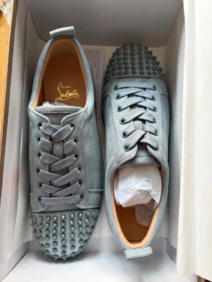 Gråa Christian Louboutin sneakers - Snygga grå sneakers från Christian Louboutin med ikoniska röda sulor och coola nitar framtill. Skorna har en låg siluett, är gjorda i mocka och har matchande grå skosnören. Perfekta för dig som vill sticka ut med en lyxig och edgy stil.