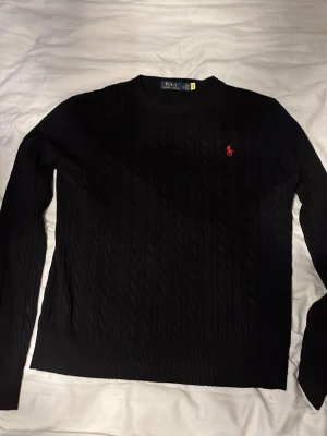 Svart stickad tröja från Polo Ralph Lauren - Kabelstickad svart tröja från Polo Ralph Lauren med rund halsringning och den klassiska röda logotypen broderad på bröstet. Långa ärmar och ribbade muddar ger en clean och tidlös look. 