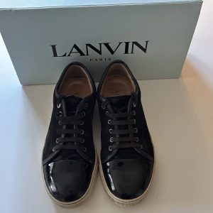 Svarta sneakers från Lanvin - Snygga svarta sneakers från Lanvin med lackad tå och detaljer i mocka. Skorna har vita sulor och klassisk snörning. Insidan är fodrad i läder för extra komfort. Perfekta för dig som gillar stilrena och exklusiva sneakers.