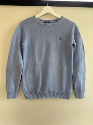 Ljusblå stickad tröja Polo Ralph Lauren - Snygg ljusblå stickad tröja från Polo Ralph Lauren med klassisk rund halsringning och diskret broderad logga på bröstet. Tröjan har ribbade muddar vid ärmslut och nederkant. Perfekt för dig som gillar stilrena och tidlösa plagg.