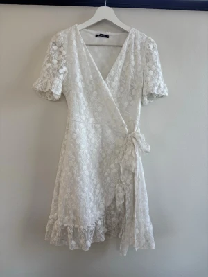 Vit klänning från Gina Tricot midsommar studenten  - Säljer en superfin vit omlottklänning från Gina Tricot med blommigt spetsmönster, volangkant nertill och knyt i midjan. Klänningen har korta ärmar och v-ringning, perfekt för sommarens alla tillfällen.