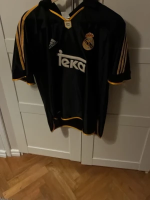 Real Madrid svart matchtröja ifrån Adidas  - Säljer en svart Real Madrid matchtröja från Adidas med gula detaljer och Teka som sponsortryck. Tröjan har klubbmärke på bröstet, klassiska Adidas-ränder på axlarna och är tillverkad i ett lätt, sportigt material. Perfekt för fotbollsträning eller att bära till vardags. Köptes i staden Madrid inne i arenan!