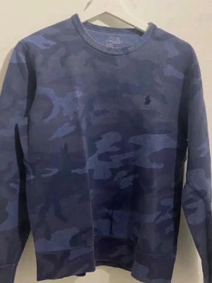 Blå camo sweatshirt från Polo Ralph Lauren - Snygg blå sweatshirt med camouflagemönster från Polo Ralph Lauren. Tröjan har rund halsringning, långa ärmar och en diskret broderad logga på bröstet. Perfekt för en avslappnad streetwear-look. Pris kan diskuteras
