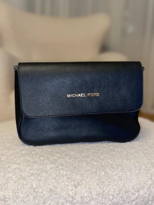 Svart axelväska -  Säljer den nu fick den som present när jag fyllde år behöver inte den länge Snygg svart axelväska med guldfärgad logga på framsidan. Väskan har ett stilrent och klassiskt design med lock och magnetknapp, samt ett innerfack med dragkedja. Materialet är skinn och den har guldfärgade detaljer.