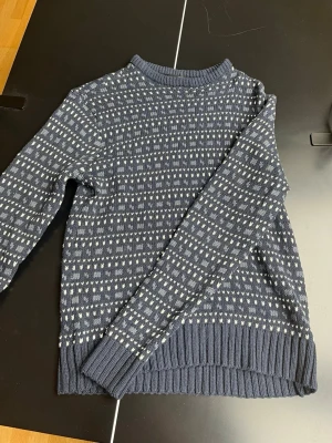 Mörkblå stickad tröja 157 - Mysig mörkblå stickad tröja från 157 med vitt mönster över hela tröjan. Tröjan har rund hals, långa ärmar och breda ribbade muddar vid ärmslut och nederkant. Perfekt för kalla dagar och enkel att matcha med jeans eller kjol.