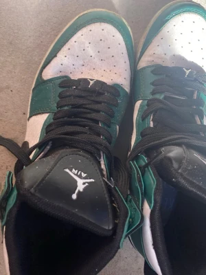 Air Jordan 1 Mid grön/vit/svart - Snygga Air Jordan 1 Mid sneakers i grönt, vitt och svart. Skorna har klassisk hög siluett, svart snörning och perforerad tåbox. Tillverkade i läder med ikoniska Jumpman-loggan på plösen. Perfekta för dig som gillar streetwear och vill sticka ut.