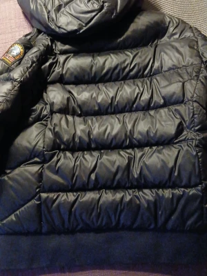 Svart pufferjacka från Parajumpers - Snygg svart pufferjacka från Parajumpers med huva och dragkedja framtill. Jackan har quiltad design, två sidofickor med dragkedja och en patch på ärmen. Perfekt för kalla vinterdagar och riktigt varm tack vare det vadderade materialet.