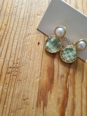 Guldörhängen med pärlor och grön sten - Snygga örhängen med rund vit pärla upptill och en större rund grön sten med marmorerat mönster nedtill. Båda delarna är infattade i guldfärgad metall. Perfekta för dig som vill ha något unikt och trendigt.