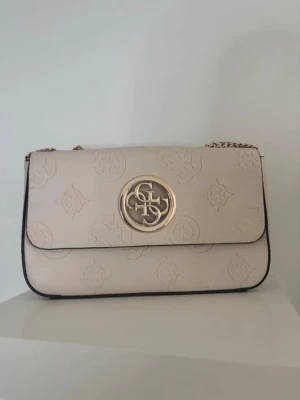 Beige handväska från Guess - Stilren beige handväska från Guess med guldfärgad logga framtill och präglat mönster över hela väskan. Kedjerem i guld och svart kant runt väskan ger en snygg kontrast. Perfekt accessoar för att lyfta din outfit.