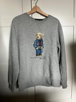 Grå sweatshirt Polo Bear Ralph Lauren - Grå sweatshirt från Ralph Lauren med ikoniskt Polo Bear-tryck framtill. Tröjan har rund halsringning, långa ärmar och en relaxed passform. Perfekt för en chill och stilren look. Nypris: 1800.