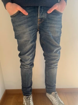 Blå Nudie Jeans  - Snygga blå slim fit jeans från Nudie Jeans med klassisk femficksmodell och coola slitningar. Jeansen har orangea kontrastsömmar, normal midja och patch med logga bak i midjan. Tillverkade i denim av bomull. Perfekta för våren! Hör av er vid frågor! 