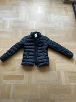 Svart pufferjacka från Tommy Hilfiger - Snygg svart pufferjacka från Tommy Hilfiger med diskret logga på bröstet. Jackan har hög krage, dragkedja framtill och två sidofickor. Tillverkad i glansigt material och perfekt för kalla vinterdagar.