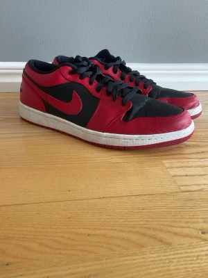 Nike Air Jordan 1 Low svart/röd - Säljer ett par Nike Air Jordan 1 Low i svart och rött läder med klassisk röd swoosh och svart snörning. Inprincip oanvända. Skorna har vit mellansula och röd yttersula. 
