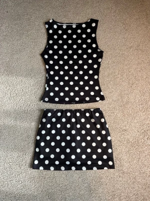 Svart prickigt set med linne och kjol - Snyggt tvådelat set med svart linne och matchande kjol, båda med stora vita prickar. Linnet är ärmlöst och figurnära, kjolen är kort och har resår i midjan. Inbyggda shorts 