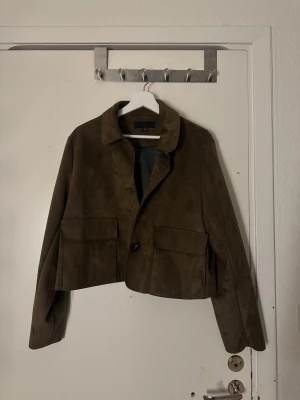 Brun croppad mockajacka - Snygg brun croppad jacka i mocka med klassisk krage och stora fickor framtill. Jackan har en knappstängning och långa ärmar, perfekt för dig som vill ha en stilren och trendig look. Jackan är i XL då den egentligen är ganska kort, men passar mig som är 165 cm lång🥰