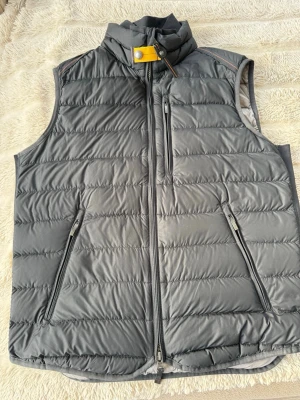 Parajumpers väst.Mörkgrå dunväst med dragkedjor - Snygg mörkgrå dunväst med ståkrage och gul detalj vid halsen. Västen har tre dragkedjeförsedda fickor framtill och quiltad design. Perfekt för lager-på-lager och håller dig varm under kyliga dagar.