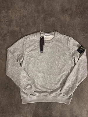 Grå sweatshirt från Stone Island - Snygg grå sweatshirt från Stone Island med klassisk rund halsringning och ribbade muddar. Tröjan har den ikoniska logotypen på vänster ärm och är tillverkad i mjukt bomullsmaterial. Perfekt för en avslappnad och stilren look.