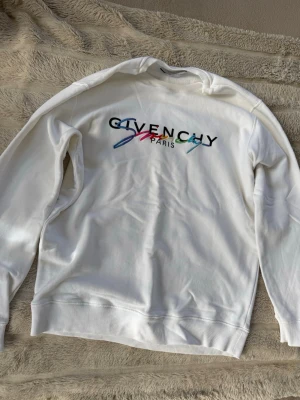 Vit sweatshirt från Givenchy Paris - Säljer en vit sweatshirt från Givenchy Paris med färgglad broderad logga på bröstet. Tröjan har rund halsringning, ribbade muddar vid ärmslut och nederkant samt en mjuk bomullskänsla. Perfekt för dig som gillar exklusiva streetwear-plagg.