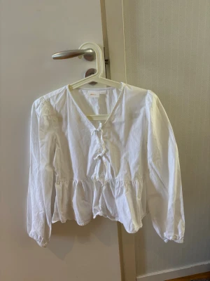 Vit blus med knytning framtill - Söt vit blus med v-ringning och knytning framtill. Blusen har långa ärmar med lätt puff och resår vid ärmslut. Den har en avslappnad passform och volang nedtill som ger en romantisk känsla. Perfekt att styla med jeans eller kjol. Storlek 146/152