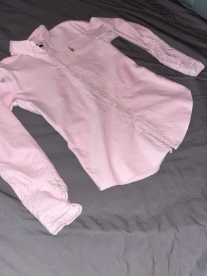 Ljusrosa skjorta från Ralph Lauren - Klassisk ljusrosa skjorta från Ralph Lauren med button-down krage och broderad logga på bröstet. Skjortan har långa ärmar, vita knappar och är tillverkad i mjuk bomull. Perfekt för en clean och stilren look. Hör av er med bud och eventuella frågor 