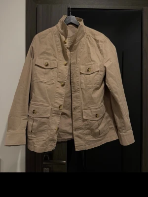 Beige Safari jacket från Oscar Jacobson - Stilren beige jacka från Oscar Jacobson med fyra framfickor. Jackan har knäppning med stora knappar och är tillverkad i bomull. Perfekt för dig som gillar klassisk och enkel design.