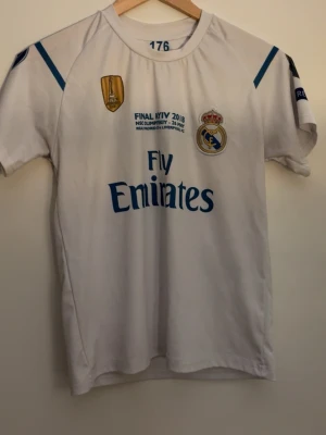 Real Madrid matchtröja 2018 - Säljer en vit Real Madrid matchtröja från Champions League-finalen 2018 med blå detaljer, klubbmärke och Fly Emirates-tryck på bröstet. Tröjan har korta ärmar och rund hals. Perfekt för dig som älskar fotboll och vill visa ditt stöd för laget.