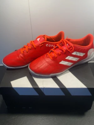 Adidas Copa röda fotbollsskor - Snygga röda Adidas Copa fotbollsskor med vita detaljer och klassiska tre ränder på sidan. Skorna har snörning och ovandel i syntetmaterial som ger bra känsla på bollen. Perfekta för dig som vill sticka ut på planen och ha grym kontroll.