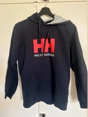 Mörkblå Helly Hansen hoodie med logga - Snygg mörkblå hoodie från Helly Hansen med stor röd HH-logga och vit text på bröstet. Grå insida på huvan och matchande svarta dragsnören. Klassisk känguruficka framtill och ribbade muddar vid ärmar och nederkant. Perfekt för en avslappnad streetwear-look. 