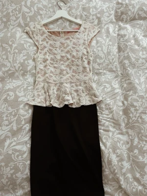 Beige spetsklänning Pernilla Wahlgren  - Snygg klänning med beige spets upptill och svart, rak kjol. Överdelen har volangkant och korta ärmar, samt ett vackert spetsmönster.