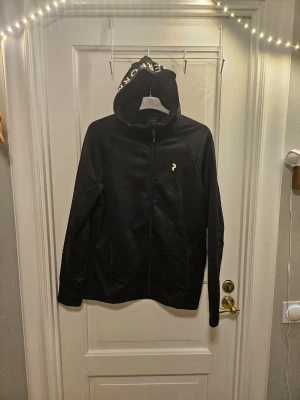Svart zip-hoodie från Peak Performance - Snygg svart hoodie från Peak Performance med dragkedja framtill och huva där det står 'PERFORMANCE' tryckt i vitt. Liten logga på bröstet. Perfekt för en sportig och avslappnad stil.  Den är nästan aldrig använd och köptes för 1600 kr. Kan köpas tillsammans med trashier tröja för 1400 kr. 