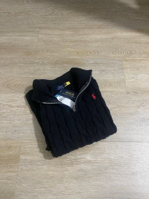 Svart kabelstickad tröja Polo Ralph Lauren - Kabelstickad svart tröja från Polo Ralph Lauren med halv dragkedja och klassisk röd logga på bröstet. Tröjan har hög krage och ribbade muddar. Perfekt för dig som gillar stilrena och tidlösa plagg. Materialet är mjukt och håller värmen.