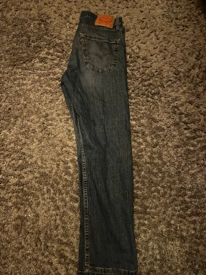 Levi's raka blå jeans - Klassiska blå jeans från Levi's med rak passform och femficksmodell. Jeansen har en mörk tvätt, tydliga sömmar och Levi's ikoniska läderpatch bak i midjan. Tillverkade i slitstark denim, perfekta för en avslappnad och tidlös stil.