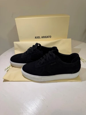  Axel Arigato skor - Stilrena svarta sneakers från Axel Arigato i mocka med vit sula. Klassisk låg modell med svarta skosnören och diskret logga på sidan. Perfekta för dig som gillar minimalistisk och clean design. Använd cirka 1-3 gånger, pris kan diskuteras