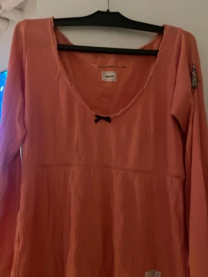 Persikofärgad topp med rosett från Impeccable - Söt långärmad topp från odd molly med v-ringning och liten svart rosett framtill. Tillverkad i ett lätt, krinklat material som ger en avslappnad känsla. Perfekt för dig som gillar feminina detaljer och mjuka färger. Tröjan är sliten och har 2 lagningar men som inte syns i första anblicken. Rosa/persilofärgad