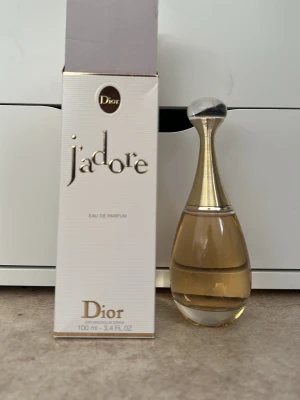J'adore Eau de Parfum från Dior - J'adore Eau de Parfum från Dior i en elegant, droppformad glasflaska med guldfärgad topp och transparent kork. Flaskan innehåller 100 ml och kommer i en stilren vit kartong med gulddetaljer. En ikonisk doft som utstrålar lyx och femininitet.