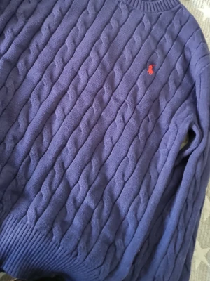 Lila kabelstickad tröja Polo Ralph Lauren - Säljer en lila kabelstickad tröja från Polo Ralph Lauren med klassisk röd logga broderad på bröstet. Tröjan har rund halsringning och ribbade muddar. Perfekt för dig som gillar tidlös och snygg stil.