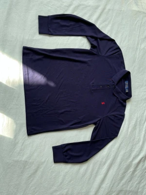 Mörkblå långärmad henley från Polo Ralph Lauren - Snygg mörkblå långärmad henley från Polo Ralph Lauren med klassisk krage och tre knappar framtill. Ikoniska röda loggan broderad på bröstet. Tillverkad i mjuk bomull som känns skön mot huden. Perfekt för en clean och stilren look.