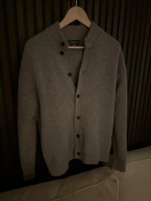 Grå kofta från Massimo Dutti - Mysig grå kofta från Massimo Dutti med ribbad krage och knäppning framtill. Tillverkad i mjukt material och har långa ärmar samt ribbade muddar. Perfekt för lager på lager och enkel att matcha med olika outfits.