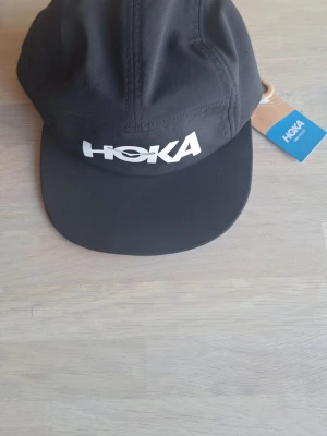 Helt ny svart keps från Hoka, one size - Snygg svart keps från Hoka med vit logga framtill. Klassisk fempanelsdesign och böjd skärm. Tillverkad i lätt och funktionellt material som passar perfekt för både träning och vardag. Justerbar baktill för optimal passform.