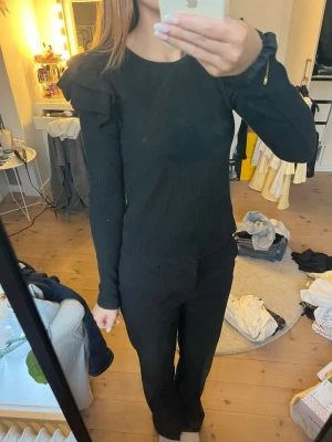 Svart ribbad topp med puffärm - Snygg svart ribbad topp med puffiga axlar och långa ärmar. Toppen har en rund halsringning och är figurnära i modellen. Perfekt att styla med både jeans och kostymbyxor för en trendig look.